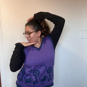 Pu&ograve; includere: Un gilet di maglia viola con scollo a V e un motivo di chiavi e volute blu scuro. Il gilet &egrave; indossato sopra una camicia a maniche lunghe nera.