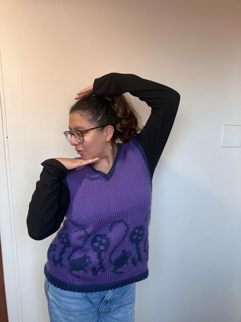 Pu&ograve; includere: Un gilet di maglia viola con scollo a V e un motivo di chiavi e volute blu scuro. Il gilet &egrave; indossato sopra una camicia a maniche lunghe nera.