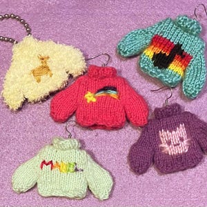 Op de afbeelding: Een verzameling miniatuur gebreide trui oorbellen en een sleutelhanger. De oorbellen zijn in verschillende kleuren, waaronder teal, roze en paars, met geborduurde ontwerpen zoals een regenboog, muzieknoot en kattengezicht. De sleutelhanger is een fluffy crèmekleurige trui met een goud geborduurd hert.