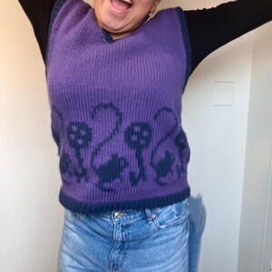 Pu&ograve; includere: Un gilet di maglia viola con un motivo a fungo blu. Il gilet ha un collo a V ed &egrave; indossato sopra una camicia a maniche lunghe nera.