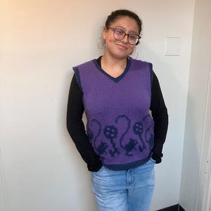 Pu&ograve; includere: Una persona indossa un gilet di maglia viola con una camicia a maniche lunghe nera sotto. Il gilet ha un collo a V e un motivo di uccelli neri sul davanti.