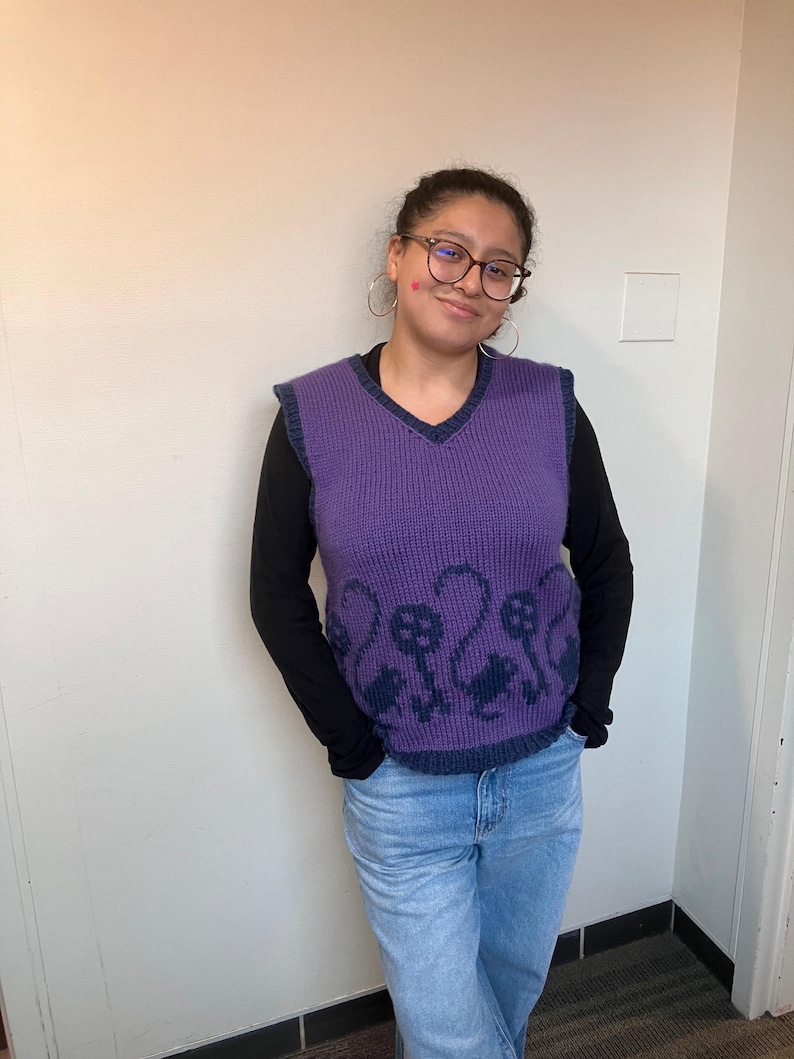 Pu&ograve; includere: Una persona indossa un gilet di maglia viola con una camicia a maniche lunghe nera sotto. Il gilet ha un collo a V e un motivo di uccelli neri sul davanti.