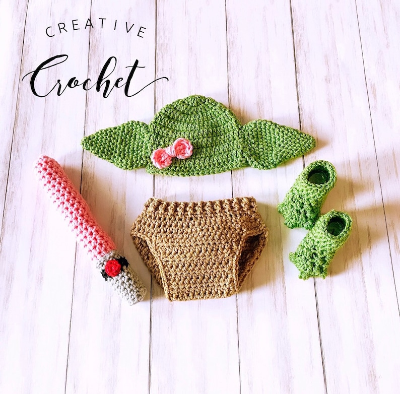 Puede incluir: Un conjunto de crochet para beb&eacute; inspirado en un personaje ficticio popular. El conjunto incluye un gorro verde con orejas, un pa&ntilde;al de tela marr&oacute;n, botines verdes y un sable de luz rosa.
