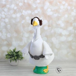 May include: A white goose figurine wearing a crocheted outfit, including a white robe, a gray belt, and a brown wig. The goose has a yellow beak and green base with gold feet. The background is a soft, blurred bokeh.