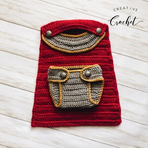 May include: A handmade red crochet baby outfit with a grey pocket and accents. The outfit features a decorative flap with two silver buttons and a matching pocket with two more buttons. The words "Creative Crochet" are in the upper right corner.