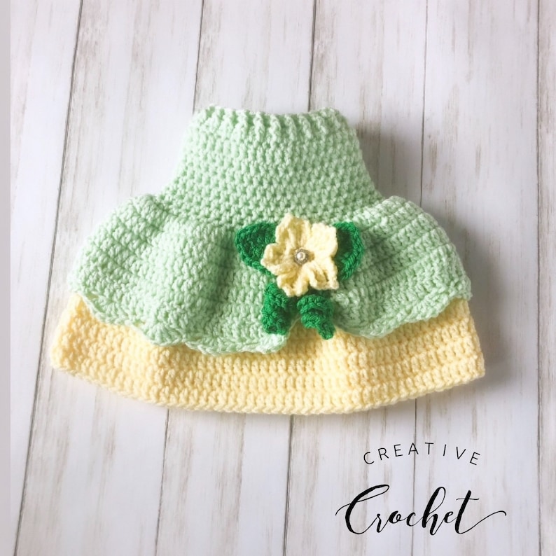 May include: A green and yellow crocheted baby hat with a flower embellishment. The hat is made with a soft yarn and has a delicate design.  The flower is made with green and yellow yarn and has a small, sparkly button in the center.  The hat is perfect for a baby girl.