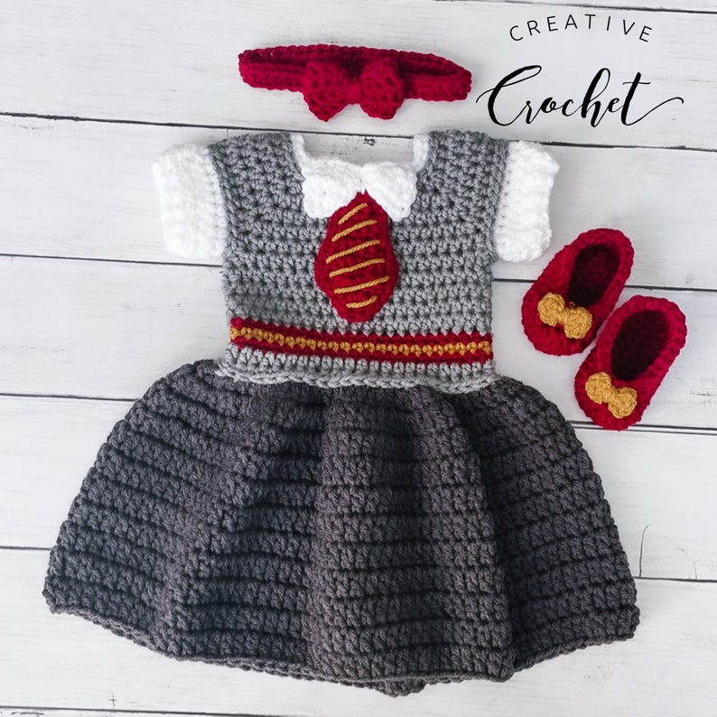 Op de afbeelding: Een grijze en witte gehaakte babyjurk met een rood strikbandje, rode schoenen met gouden strikjes en een gouden en rode stropdas. De jurk heeft een gouden en rode rand rond de taille. De tekst "CREATIVE Crochet" staat in de rechterbovenhoek van de afbeelding.
