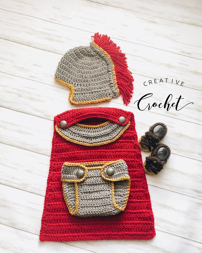 May include: A handmade crochet baby outfit in a Roman soldier theme. The set includes a grey helmet with a red crest, a red tunic with grey and gold accents, and brown sandals. The text "Creative Crochet" is also visible.
