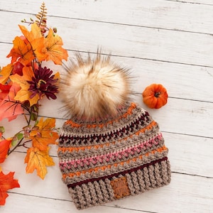 May include: A crocheted beanie with a large faux fur pom-pom on top. The hat features horizontal stripes in shades of beige, orange, pink, and dark maroon. A rectangular brown leather tag is attached to the ribbed brim with the text Sweater Weather.