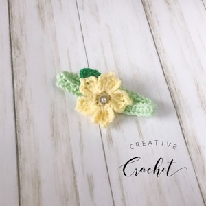 May include: A yellow crocheted flower with a pearl center, attached to a green crocheted headband. The headband is on a white wooden surface. The text "CREATIVE Crochet" is visible below the headband.