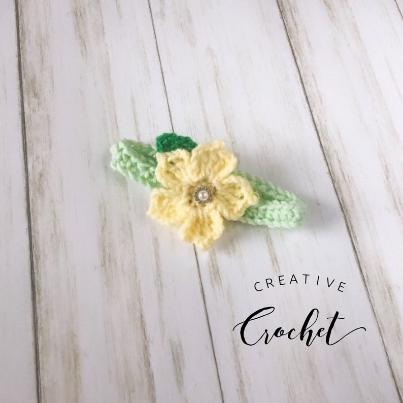 May include: A yellow crocheted flower with a pearl center, attached to a green crocheted headband. The headband is on a white wooden surface. The text "CREATIVE Crochet" is visible below the headband.