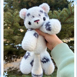 Op de afbeelding: Een handgemaakt wit en grijs gehaakt sneeuwluipaard knuffel. De knuffel heeft donkergrijze vlekken, een roze neus en zwarte ogen. De tekst "Snow Leopard Pattern" wordt in de afbeelding weergegeven.