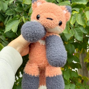 May include: A handmade plush fox toy in shades of orange, grey, and white. The fox features large, round grey paws and ears, with a white chest and belly. The toy is set against a backdrop of green leaves.