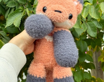May include: A handmade plush fox toy with orange, gray, and white accents. The fox has large, round gray paws and ears, with a white chest and belly. The toy is held against a backdrop of green leaves.