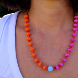 Può includere: Una collana caratterizzata da una fila di perline arancioni su un lato e perline rosa sull'altro, collegate da una singola perlina azzurra al centro. Le perline sono infilate su un sottile cordino rosso, creando un accessorio vivace e colorato.