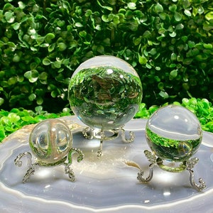 May include: Three clear crystal spheres of varying sizes on silver stands, displayed on a gray agate slice with a brown edge. The spheres are arranged in a triangular formation.