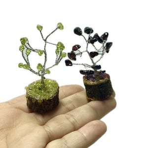 May include: Two small wire wrapped gemstone trees. The tree on the left is made with green peridot stones and the tree on the right is made with red garnet stones. Both trees are sitting on a small wooden base.