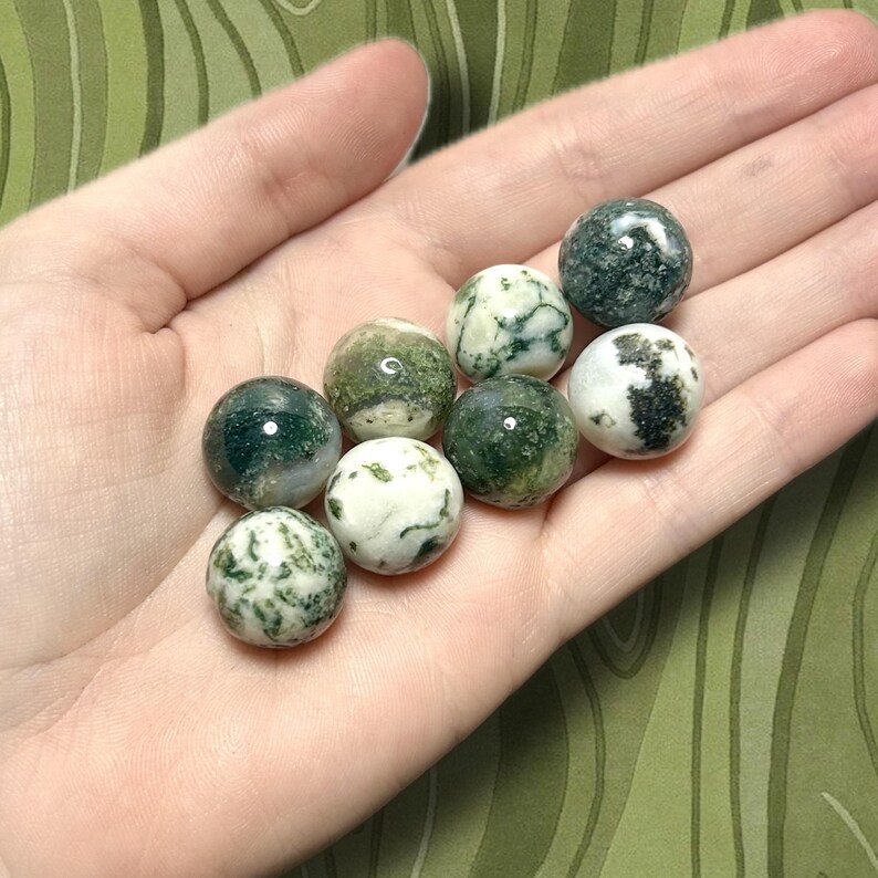 May include: A collection of eight polished moss agate spheres, each approximately 1 inch in diameter, displayed in a hand. The spheres feature a mix of translucent white and various shades of green, with unique patterns. The background is a green, patterned fabric.
