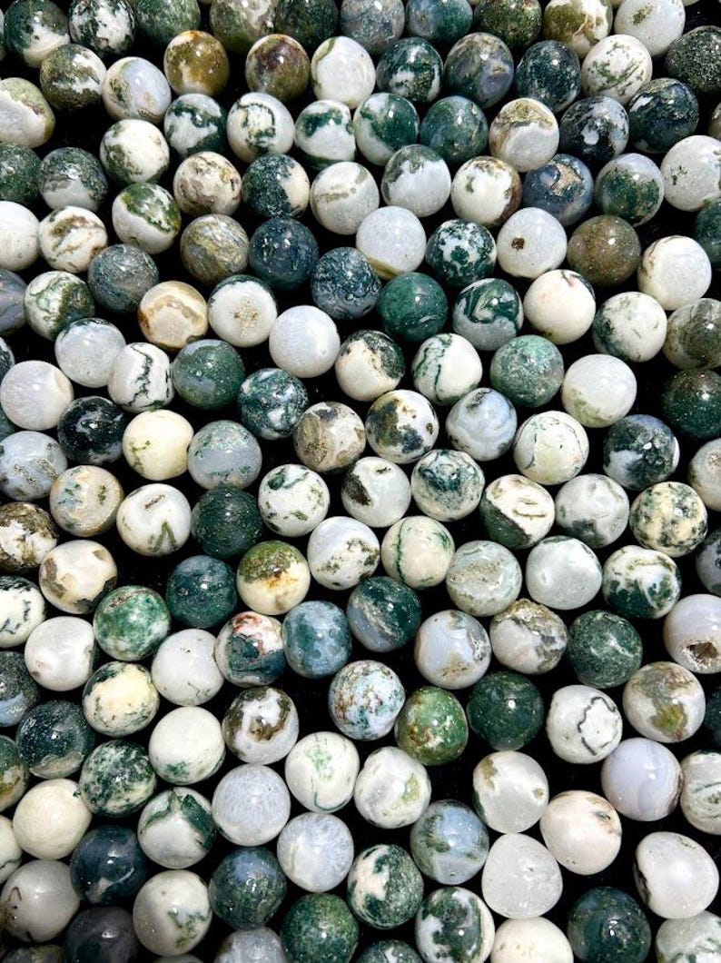May include: A collection of round, polished moss agate gemstones. The stones vary in color, with shades of white, green, and brown. The gemstones are smooth and have unique patterns, creating a natural and earthy aesthetic.