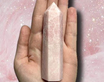 May include: A light pink crystal obelisk with a pointed top. The crystal has a textured surface with white veining. The obelisk is held in an open hand against a pink background.