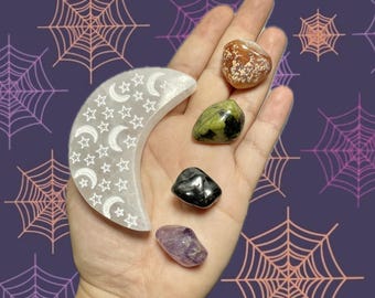 May include: A hand displays a white selenite crystal crescent moon, decorated with etched stars and moons, and several polished gemstones. These include a speckled brown and white stone, a green stone, a black stone, and a purple amethyst. The background has a purple spiderweb design.