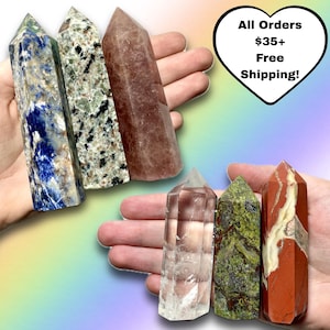 May include: Assortment of polished crystal towers in various colors, including blue, green, pink, clear, and red. The crystals are held in a hand, showcasing their unique textures and patterns. A heart-shaped graphic with text is also present.