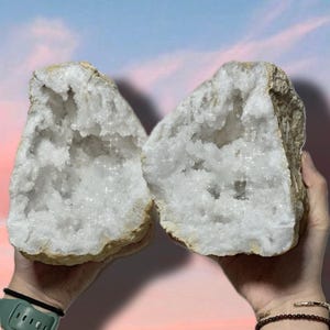 May include: Two large white crystal geode halves with a rough exterior and a sparkling, white, crystalline interior. The geode halves are being held by hands.