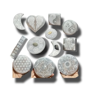 May include: A collection of 12 white selenite charging plates with various etched designs, including a heart with wings, a crescent moon, a yin yang symbol, a hamsa hand, a flower of life, and an om symbol.