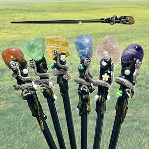 May include: A collection of handcrafted wands with dark wooden shafts. Each wand features a unique gemstone top and a silver snake design. The wands are displayed against a blurred green background, suggesting an outdoor setting.