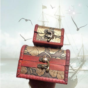 May include: Two decorative wooden treasure chests, one stacked on top of the other. The chests are red and gold, with ornate floral designs and metal clasps. The background features a ship and seagulls.