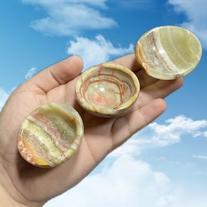 May include: Three small, round onyx bowls with a polished finish. The bowls display a natural color palette of green, cream, and reddish-brown, with unique banded patterns. The bowls are held in a hand against a blue sky background.