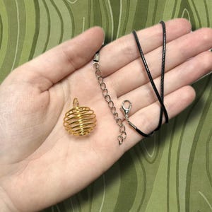May include: A gold-colored wire cage pendant necklace with a black cord. The pendant is spherical, with a spiral design. The necklace includes a silver-colored chain and clasp. The background is a green, patterned surface.