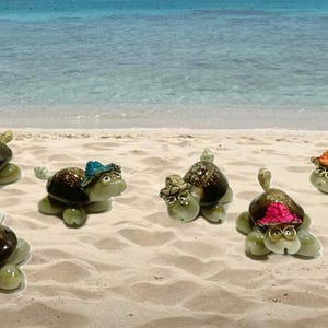 May include: Six small turtle figurines made of seashells, each wearing a different colored hat. The turtles are on a sandy beach.