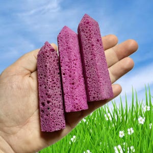 May include: Three magenta-colored, porous, obelisk-shaped objects are held in a hand against a backdrop of blue sky and green grass. The objects have a textured surface and a pointed top. The obelisks vary slightly in size.