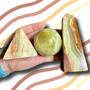 May include: A hand holds three polished stone objects: a triangle, a sphere, and a tall, rectangular piece. The triangle and rectangular piece have a cream and orange striped pattern. The sphere is a translucent green color.