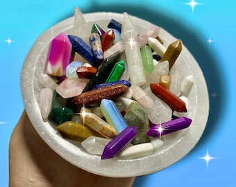 May include: A collection of colorful crystals in a white bowl. The crystals are various shapes and sizes, including pink, purple, blue, green, and clear. The bowl is held in a hand against a blue background with white star accents.