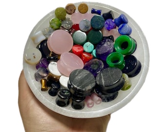 May include: A bowl filled with a variety of colourful gemstone and stone ear gauges. The gauges are in different sizes and shapes, and some have a polished finish.