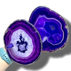 May include: Two purple agate slices with white and dark purple banding. The agate slices are round and have a polished finish.