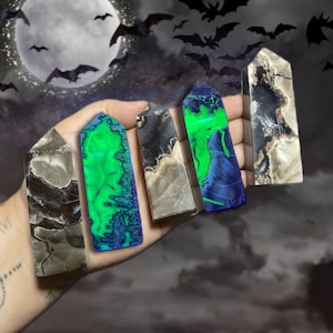 May include: Five polished gemstone obelisks in varying colors, including green, blue, black, and white, are held in a hand. The background features a full moon, bats, and a dark, cloudy sky, creating a Halloween theme.