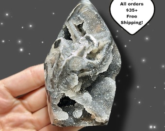 Puede incluir: Una geoda de cristal gris y blanco con una parte superior puntiaguda. El cristal tiene una superficie rugosa y texturizada con áreas de cristales brillantes. La geoda se sostiene en una mano sobre un fondo oscuro.