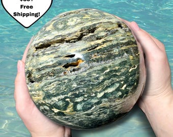 Puede incluir: Una piedra grande y esférica con un patrón en capas de tonos verdes, beige y marrones. La piedra tiene grietas naturales y una superficie pulida. La imagen incluye texto en un gráfico en forma de corazón que dice "All Orders $35+ Free Shipping!"