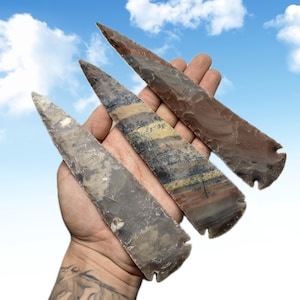 May include: Three stone arrowheads in shades of brown, gray, and tan are held in a hand against a blue sky background. The arrowheads have sharp points and notched bases, showcasing natural patterns and textures.