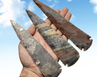 Könnte beinhalten: Drei Steinpfeilspitzen in Braun-, Grau- und Beigetönen werden in einer Hand vor blauem Himmel gehalten. Die Pfeilspitzen haben scharfe Spitzen und gekerbte Basen, die natürliche Muster und Texturen zeigen.