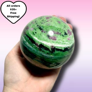 May include: A green and pink gemstone sphere. The sphere is held in a hand and has a polished surface. The stone features a mix of green, pink, and white colors, with a marbled appearance. The sphere is a decorative item.