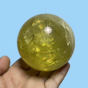 May include: A translucent, yellow-green crystal ball. The sphere is held in a hand against a light blue background. The crystal has internal fractures and a bright, reflective surface.