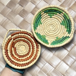 May include: Two round woven baskets. One basket has a handle and a spiral design in shades of brown and tan. The other basket has a green and tan spiral design. Both baskets are on a woven surface.