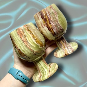 May include: Two hand-carved onyx wine glasses. The glasses feature a unique pattern of green, brown, and orange hues. The stem and base are also carved from the same stone. The glasses are held in a hand.