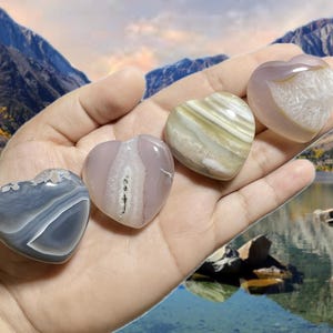 May include: Four heart-shaped stones in shades of gray, pink, and beige are held in a hand. The stones have unique patterns and textures. The background features a scenic view of mountains and a lake.