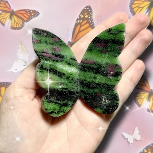 May include: A green and black butterfly-shaped stone with red flecks is held in a hand. The stone has a polished surface and a natural, organic appearance. The background features butterflies in various colors.