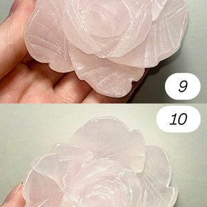 May include: A carved rose-shaped sculpture in a soft pink hue. The petals are detailed with delicate lines, creating a realistic floral design. The sculpture is held in a hand, showcasing its size and texture.
